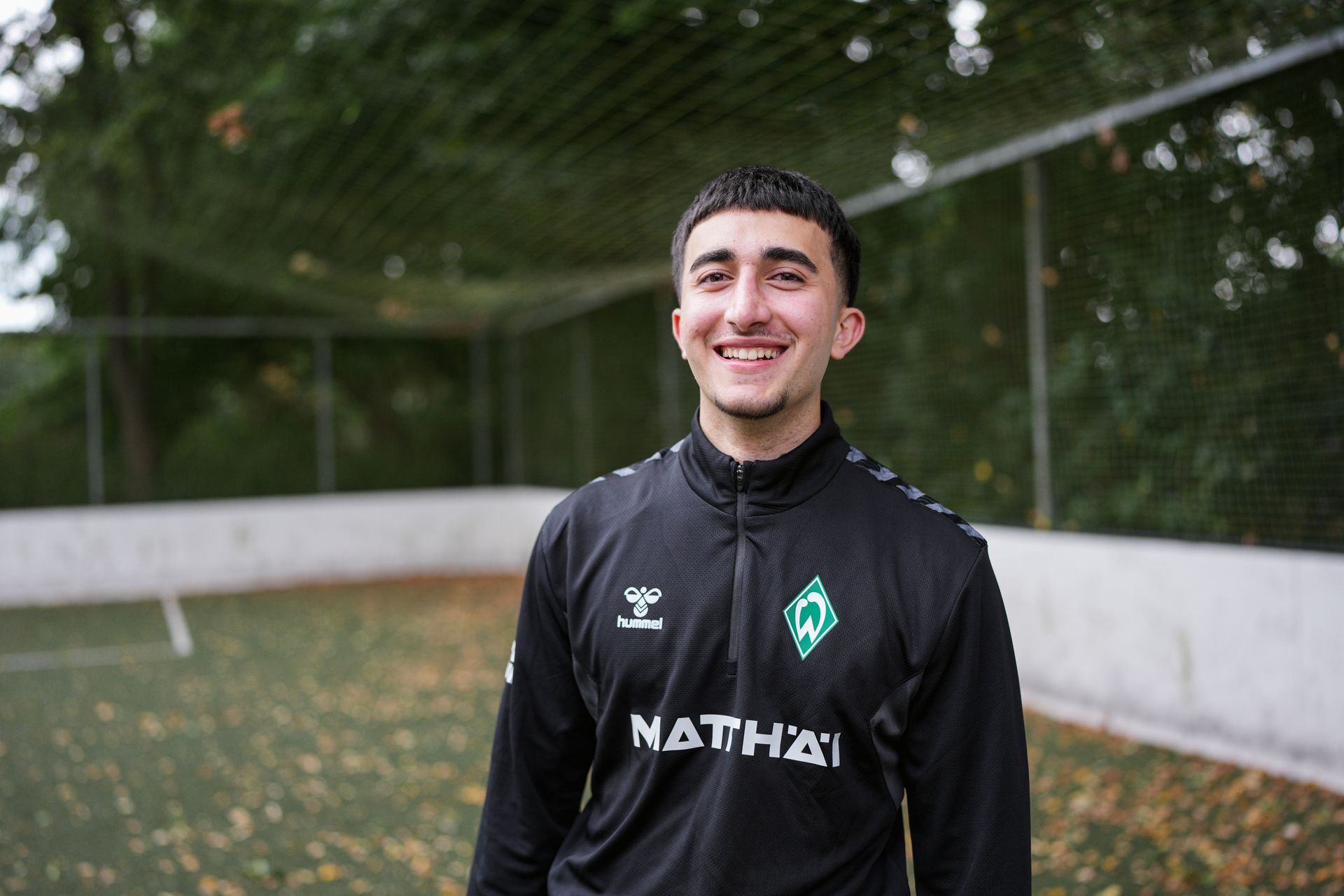 哈桑·胡鲁站在足球场上，对着镜头微笑。